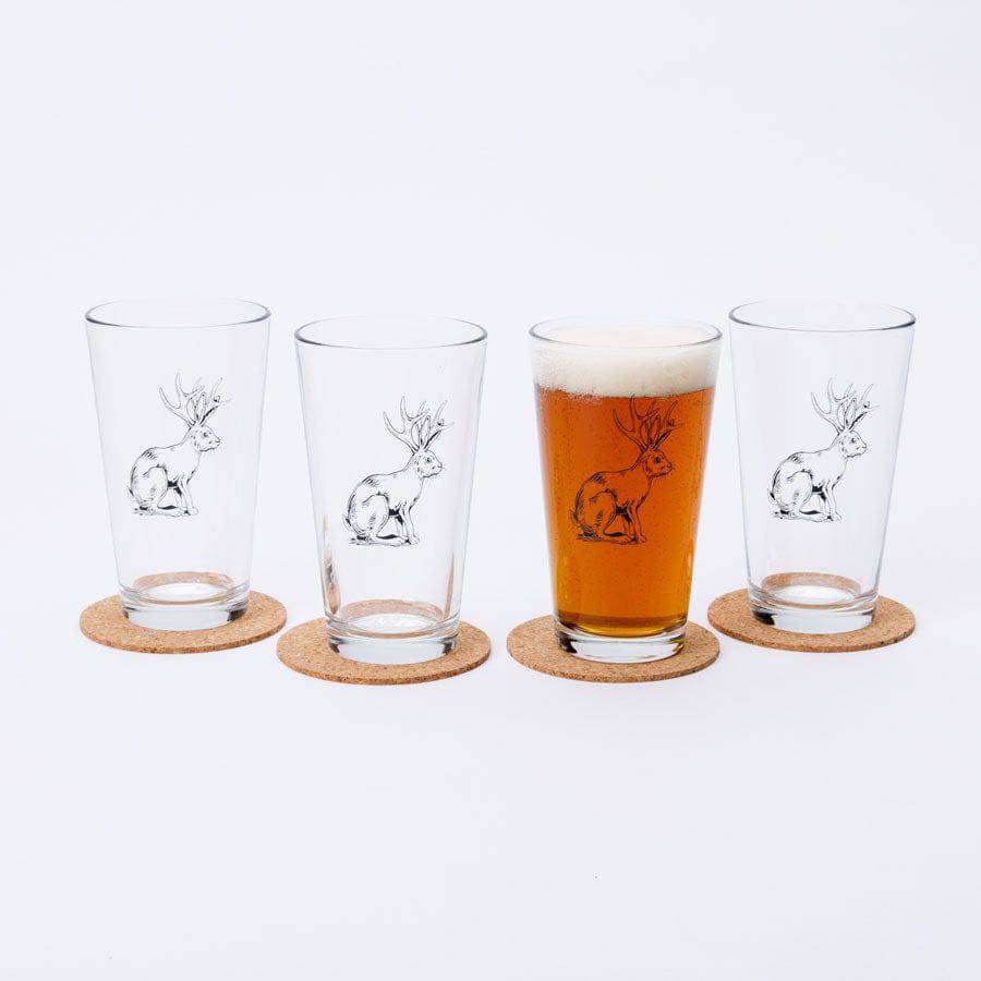 Set of four pint glasses with jackalope illustration, one filled with beer, on cork coasters