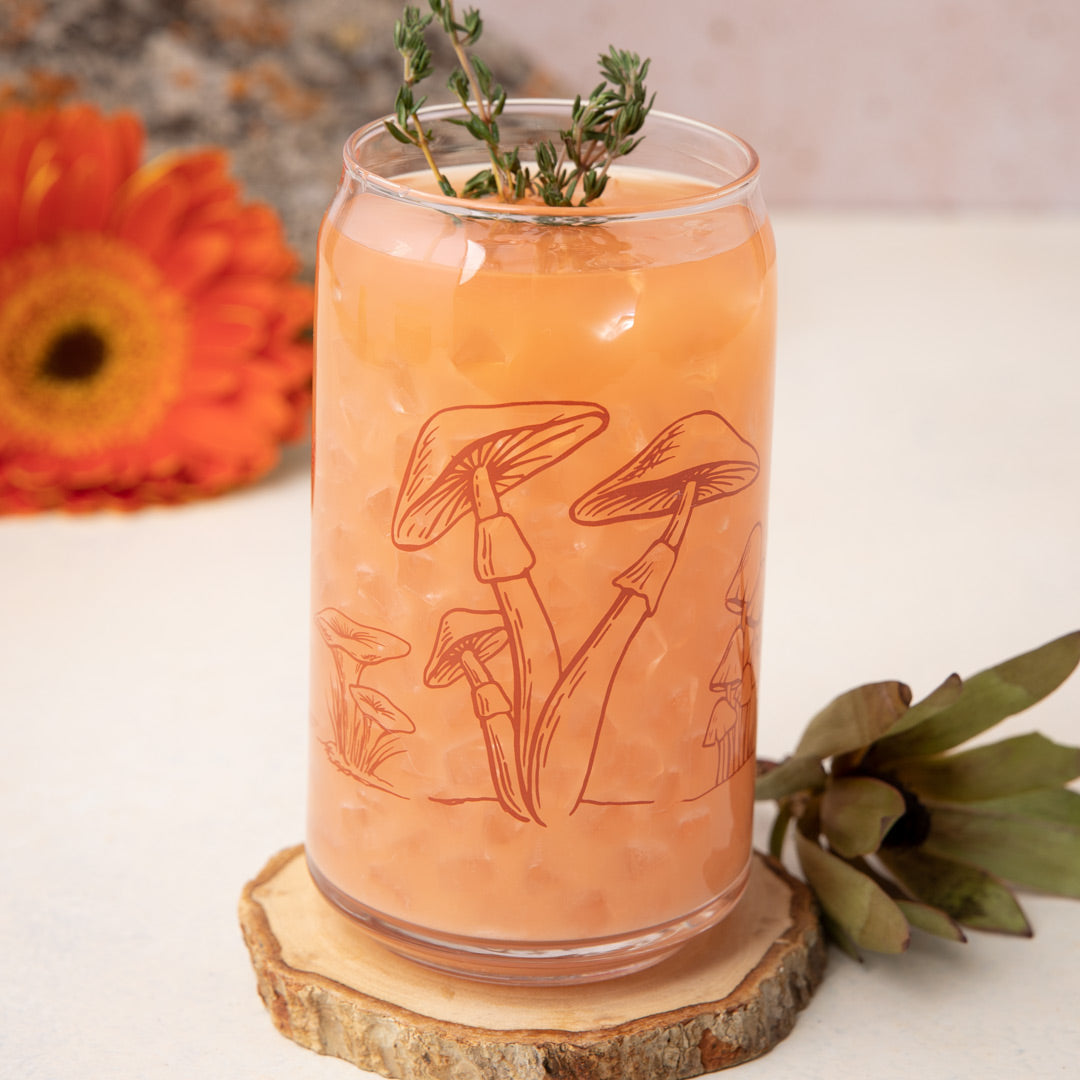 Mushroom print can-shaped glass with iced orange drink and herbs, on a wood coaster