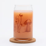 Can-shaped glass with mushroom illustrations, filled with orange beverage on cork coaster