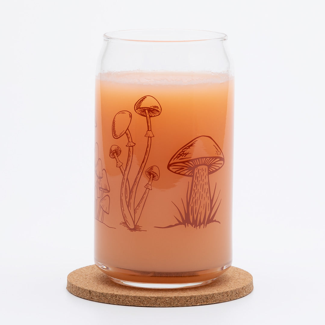 Can-shaped glass with mushroom illustrations, filled with orange beverage on cork coaster