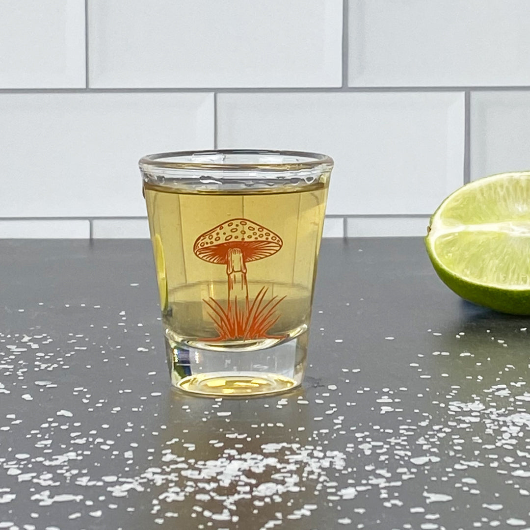 Clear shot glass with orange mushroom design, filled with liquor, lime and salt on counter.
