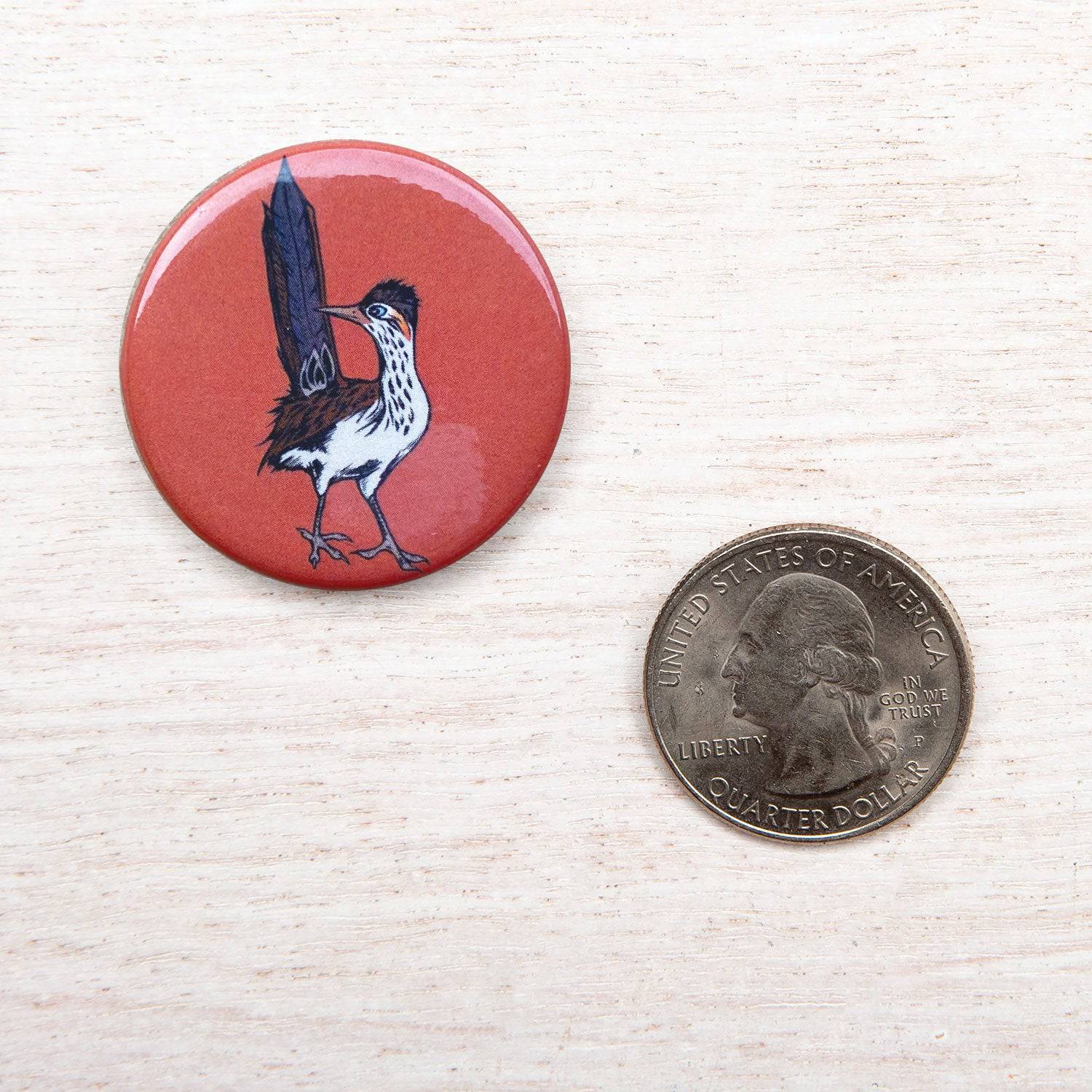 Roadrunner illustrated button pin on wood surface beside US quarter for size reference