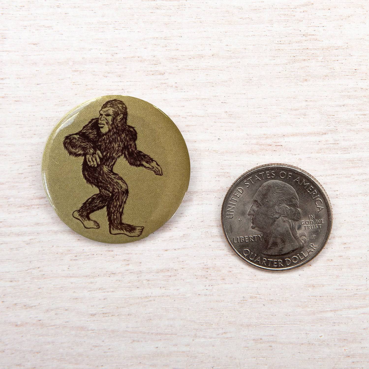 Sasquatch illustration pinback button next to a US quarter on light wood surface