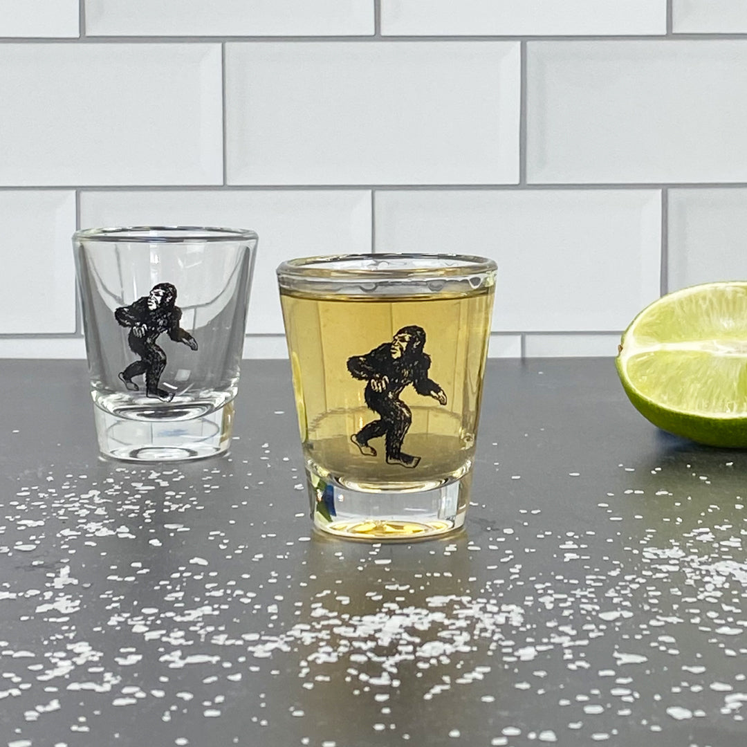 Sasquatch shot glasses with illustrated Bigfoot design, filled and empty, on a bar counter with lime and salt