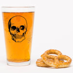 Pint glass with skull graphic filled with beer, next to two salted pretzels on white background