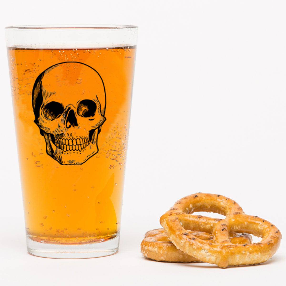 Pint glass with skull graphic filled with beer, next to two salted pretzels on white background