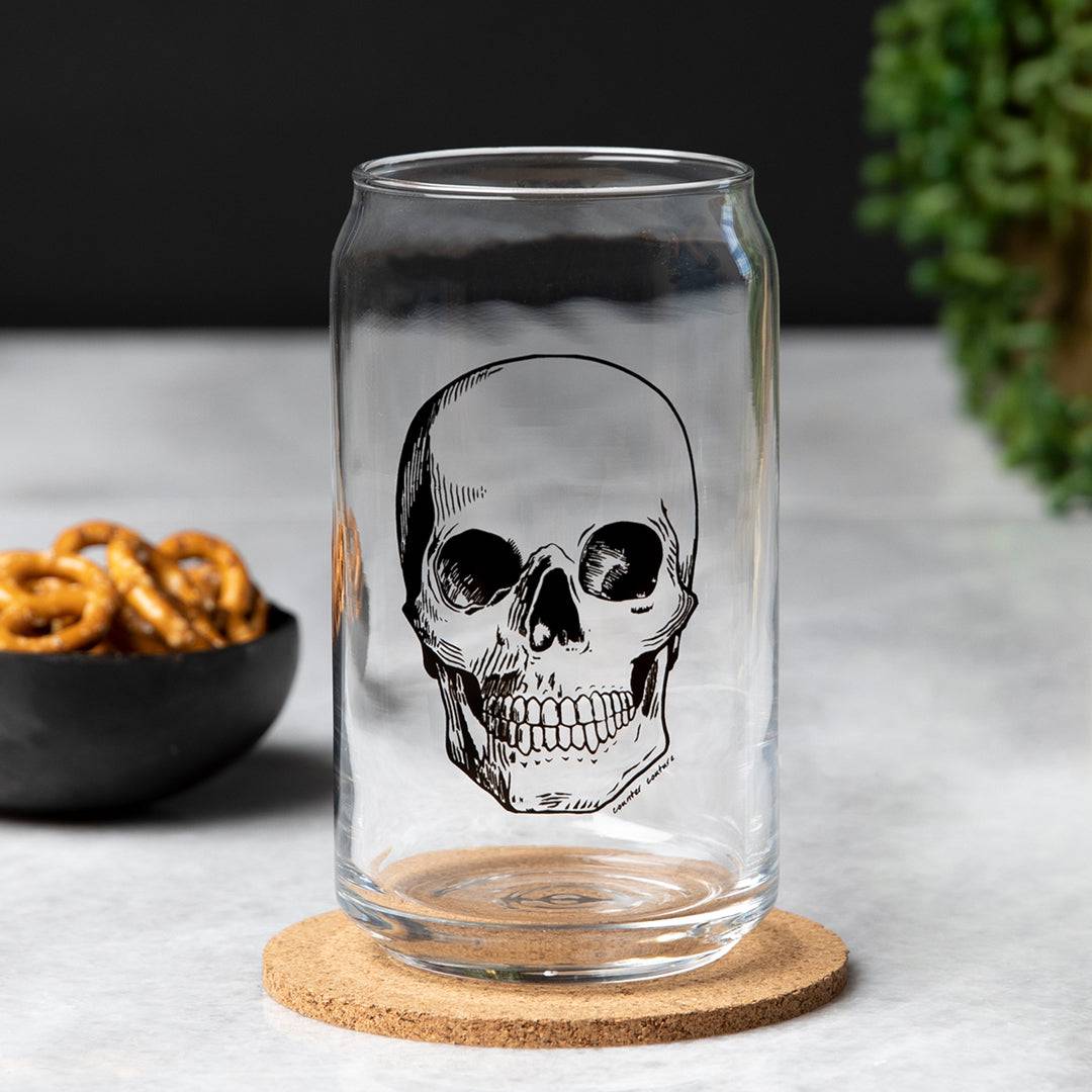 Clear can-shaped beer glass with black skull design on cork coaster, snack bowl in background