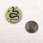 Round pin button with black snake and stars design next to a US quarter on light wood.