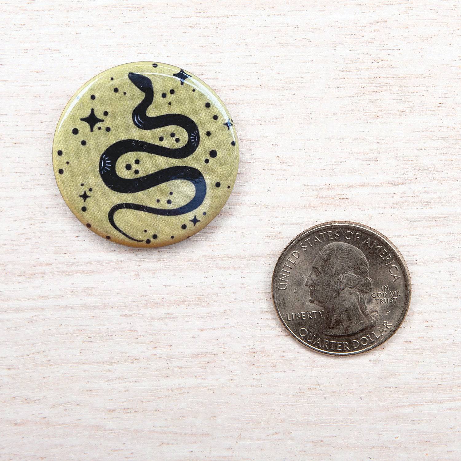 Round pin button with black snake and stars design next to a US quarter on light wood.