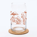 Clear can-shaped glass with brown mushroom illustration, placed on cork coaster